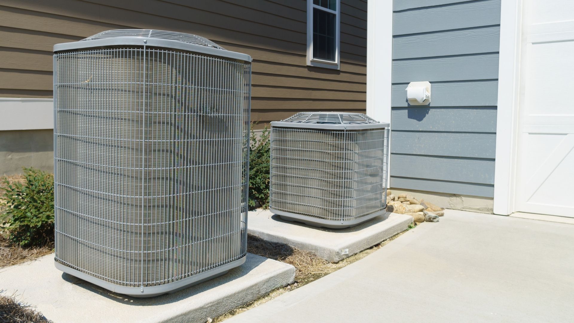Two outdoor air conditioning units beside a house and concrete driveway