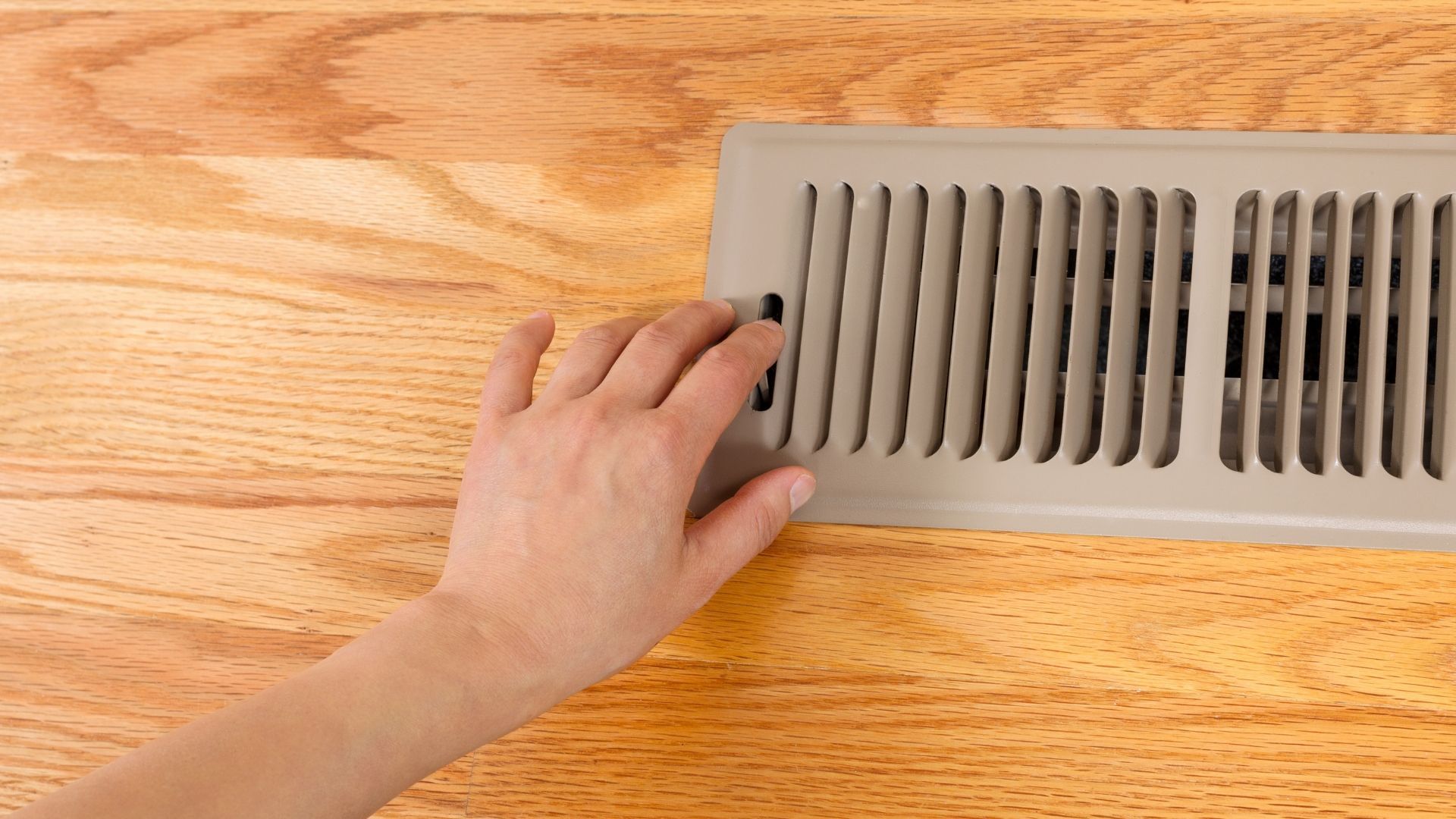 Hand reaching toward a beige floor vent on a wooden floor