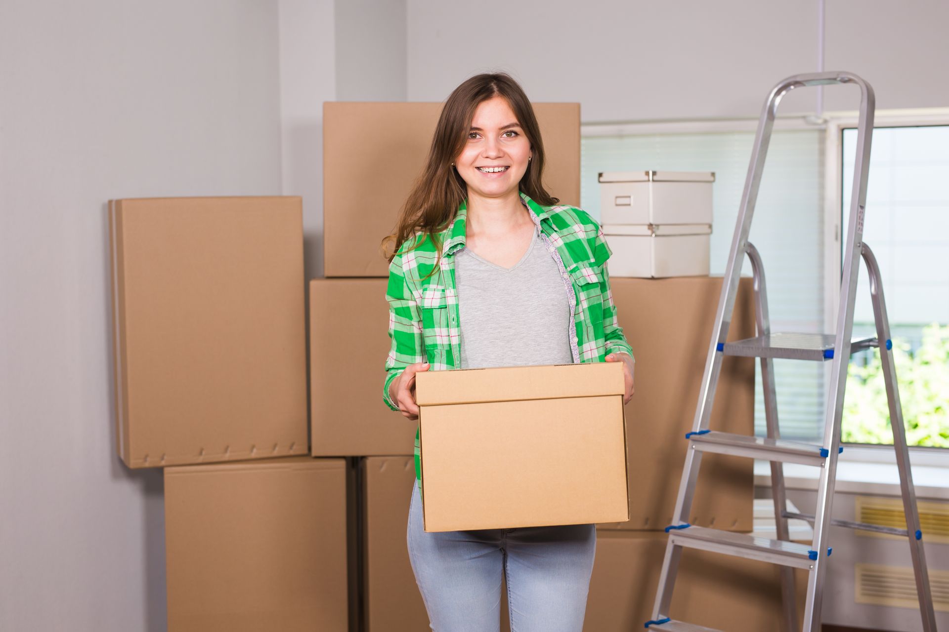 Woman holding a box, smiling in a room with moving boxes and a ladder.