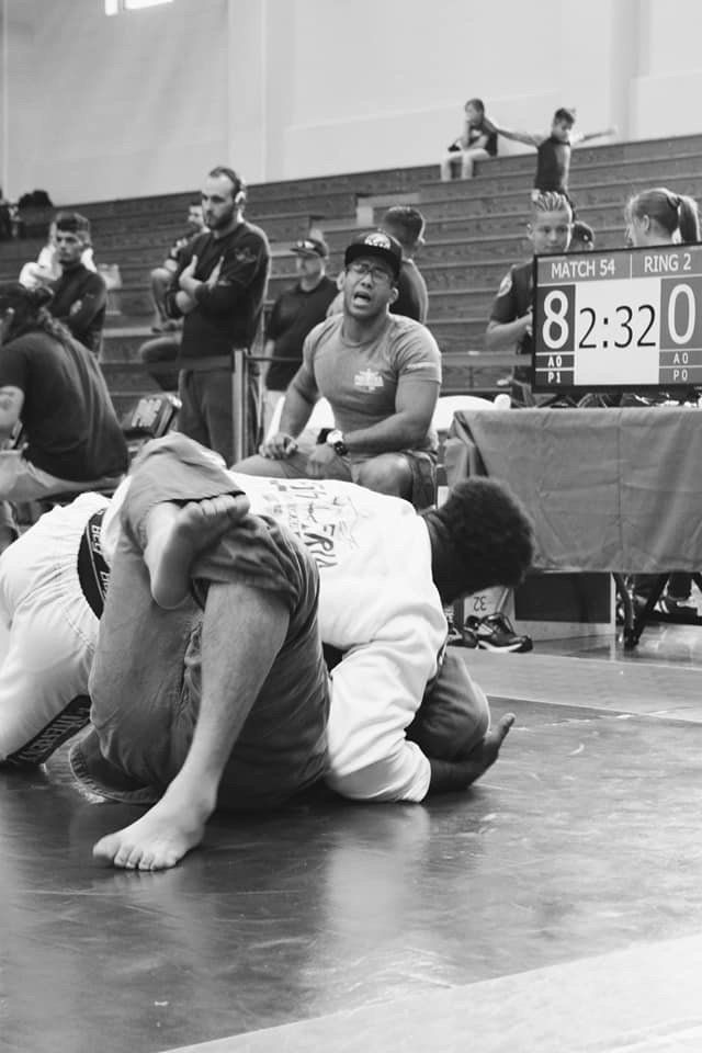 Two men grappling on a mat during a martial arts competition, with spectators and a scoreboard in the background.