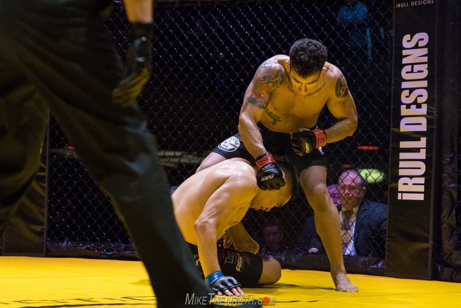 Two MMA fighters in a cage; one standing over the other on the ground, striking.