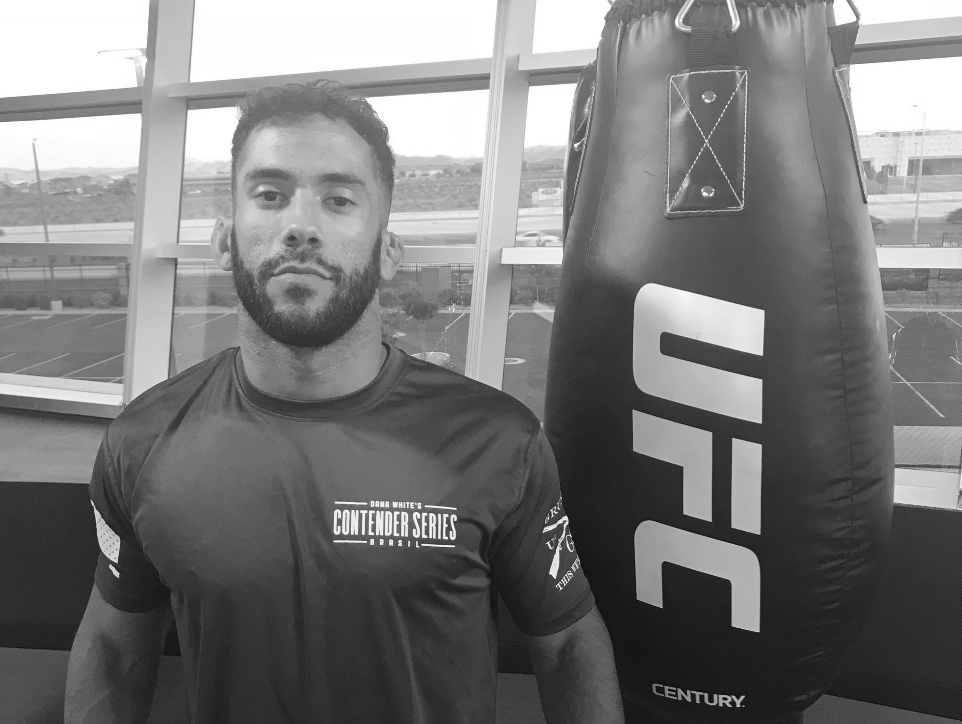 Man in a gym, posing next to a UFC punching bag. He wears a dark shirt, and the background shows a window.