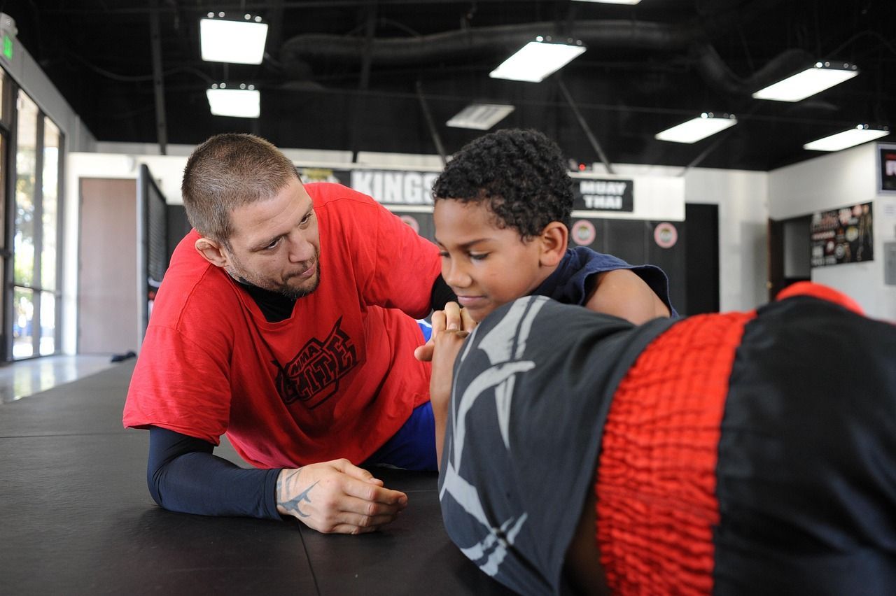 Man in red shirt coaches a boy in a gym. Both lean over another person on the mat, reviewing moves.