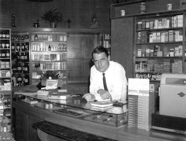 Ein Mann in Anzug und Krawatte steht hinter einem Tresen in einer Apotheke.
