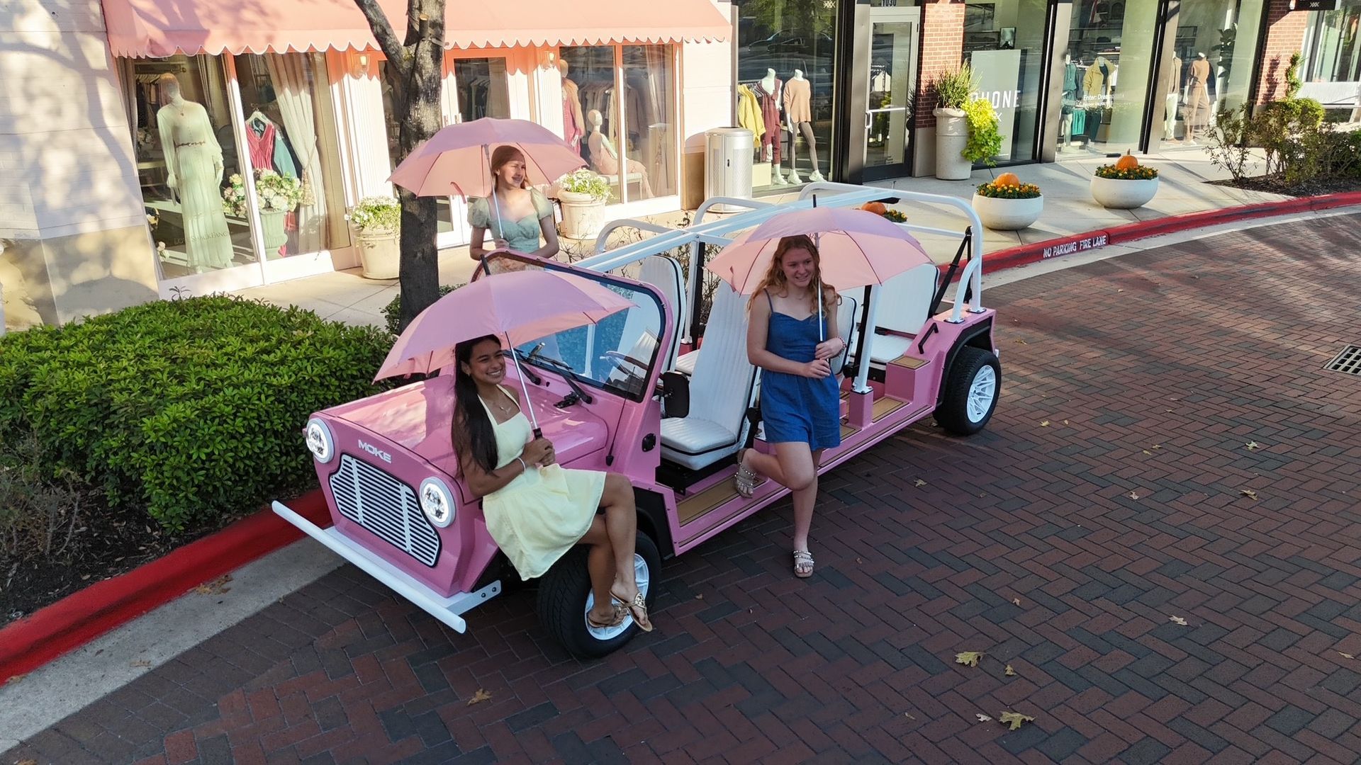 Three women with pink parasols stand near a pink golf cart, in front of shops.