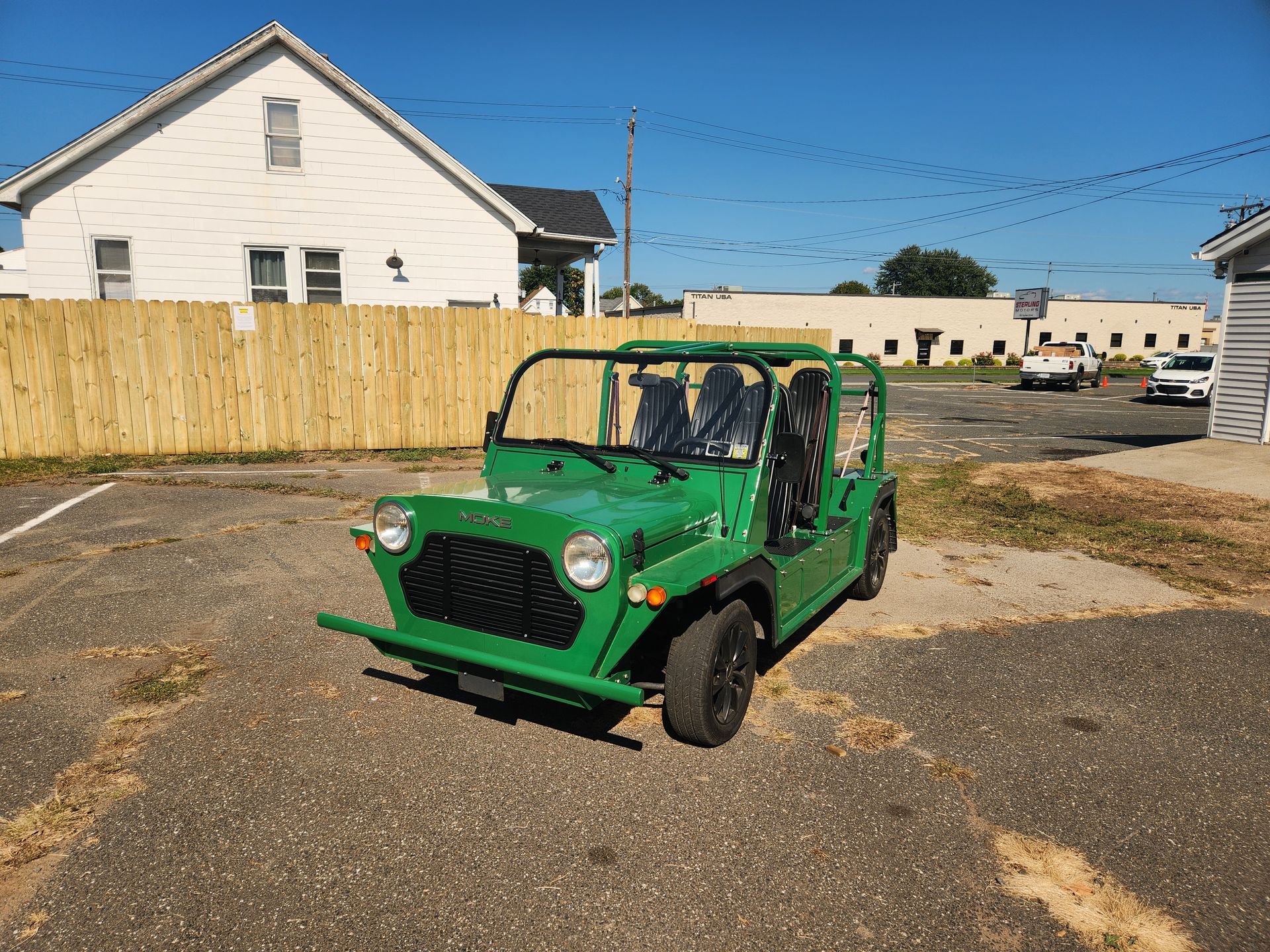 Green open-air vehicle with black seats parked on a paved lot next to a white and brown building on a sunny day.