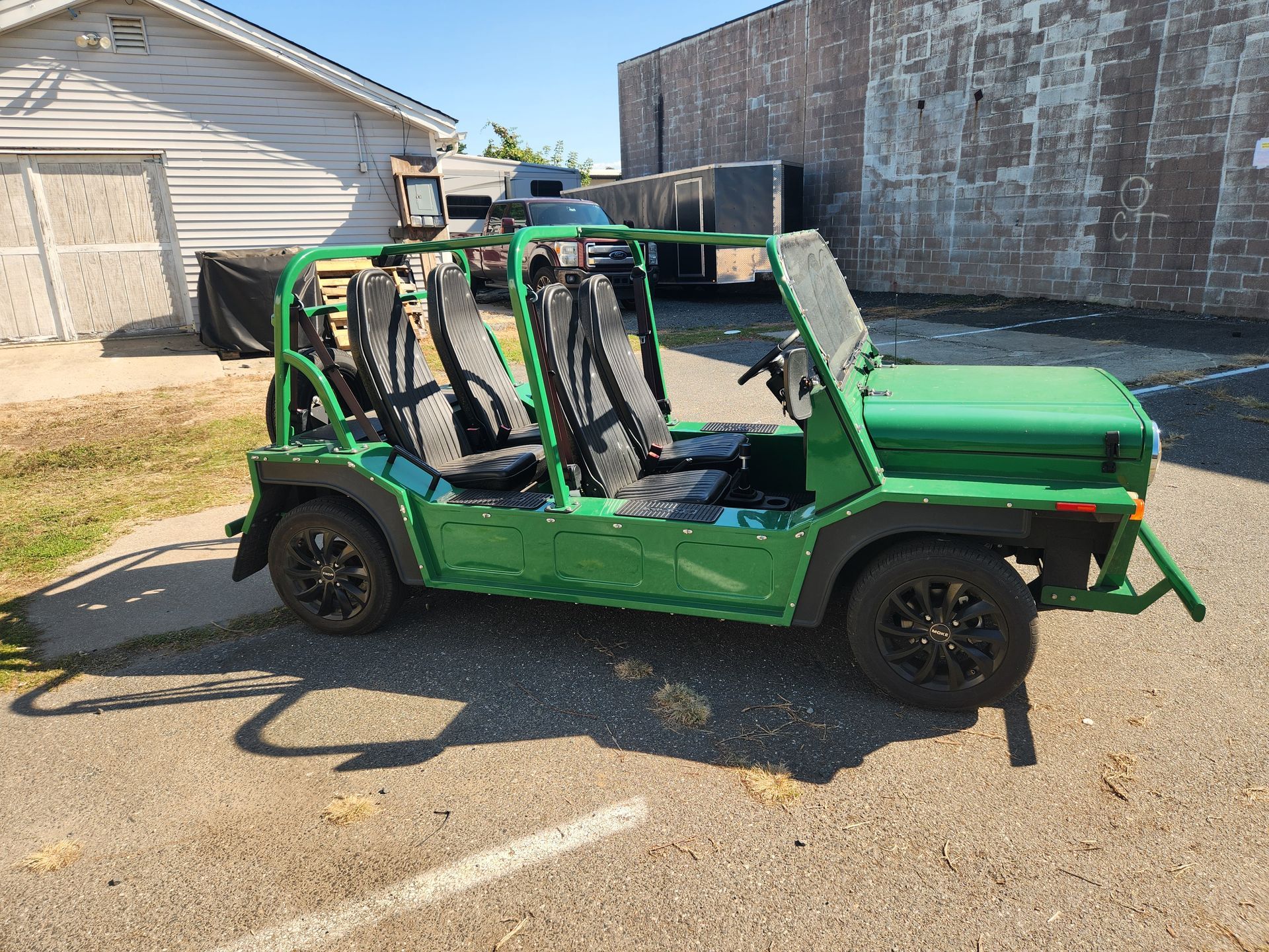 Green open-air vehicle with black seats parked on a paved lot next to a white and brown building on a sunny day.