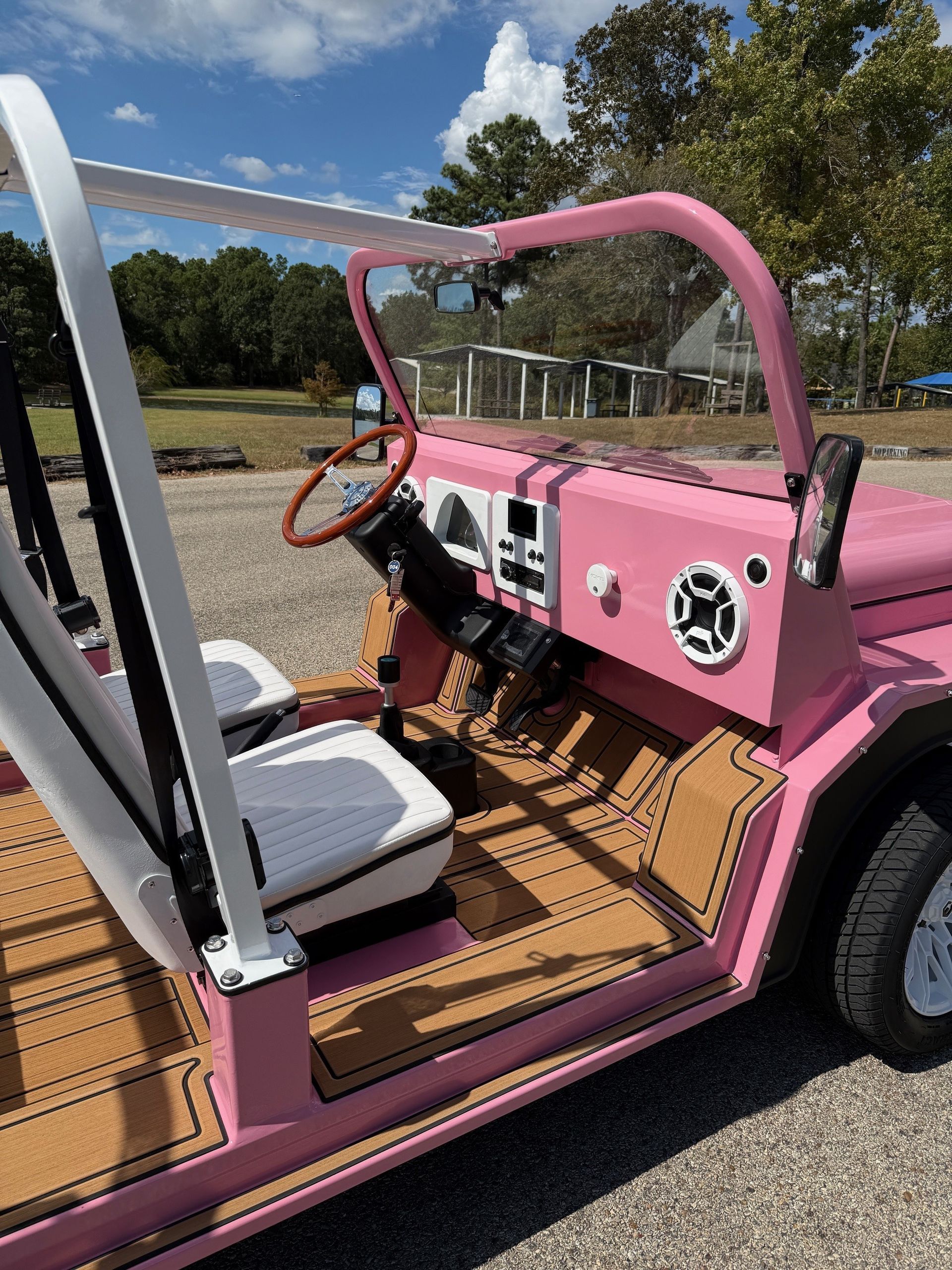 Pink and white custom open-top vehicle with six seats, parked on gravel near water and trees.