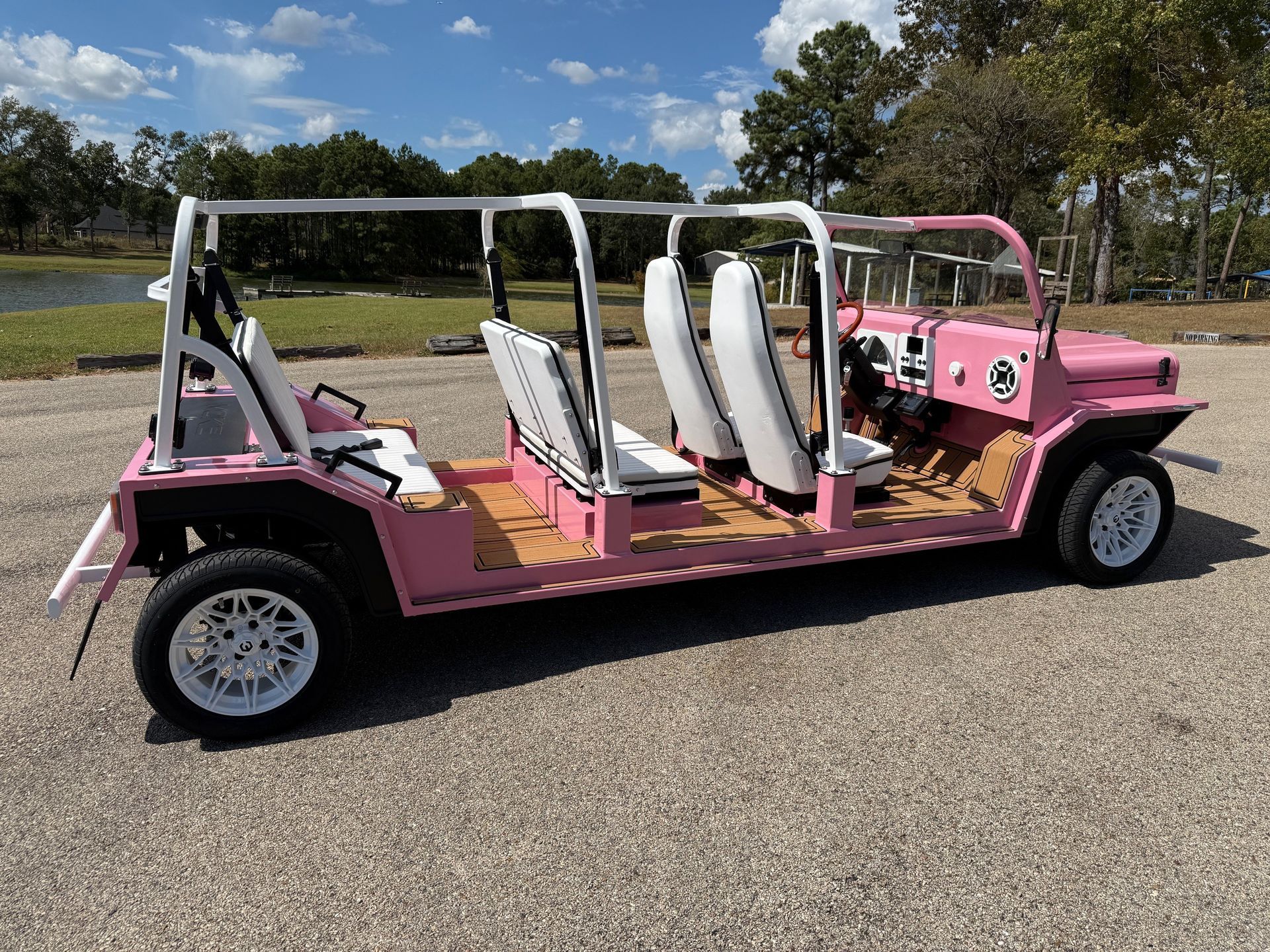 Pink and white custom open-top vehicle with six seats, parked on gravel near water and trees.