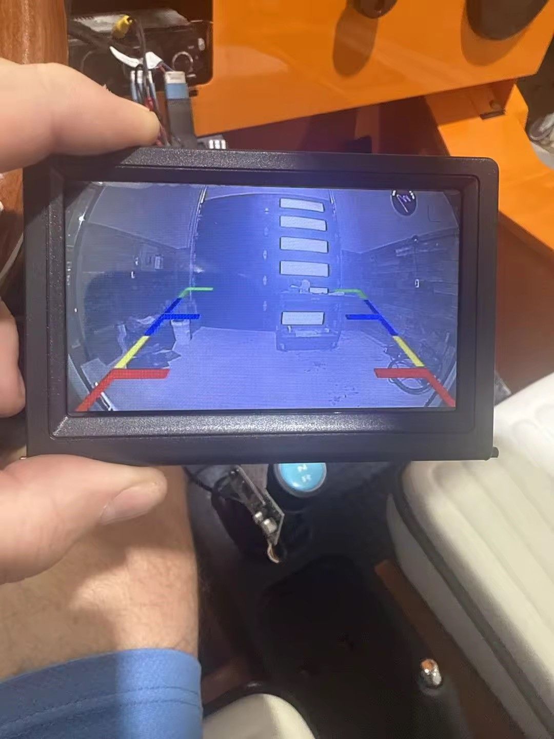 Hand holding a small screen showing a backup camera view with colored guide lines.
