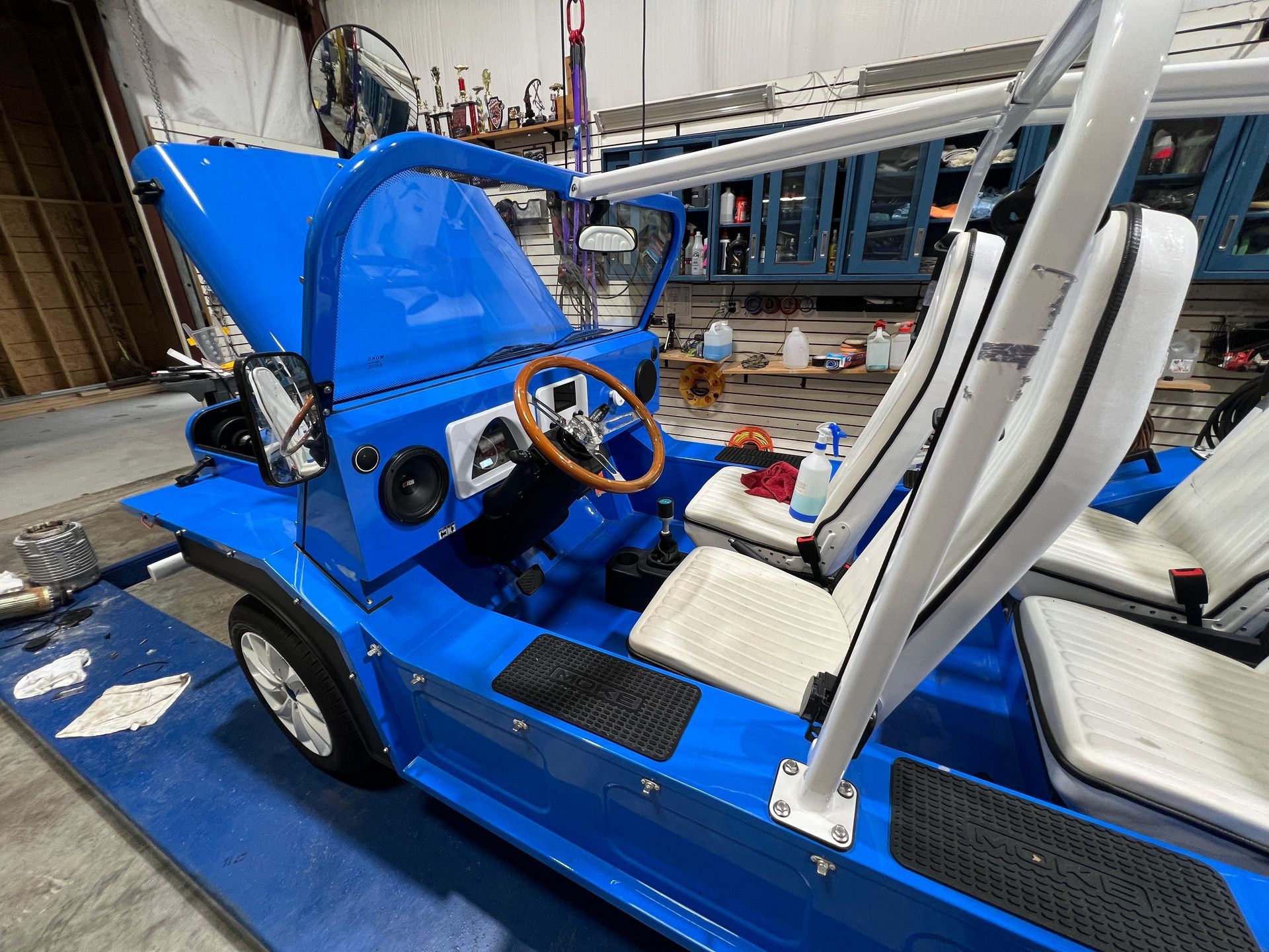 Bright blue Moke-style vehicle interior with white seats, a wooden steering wheel, and a partially open hood.