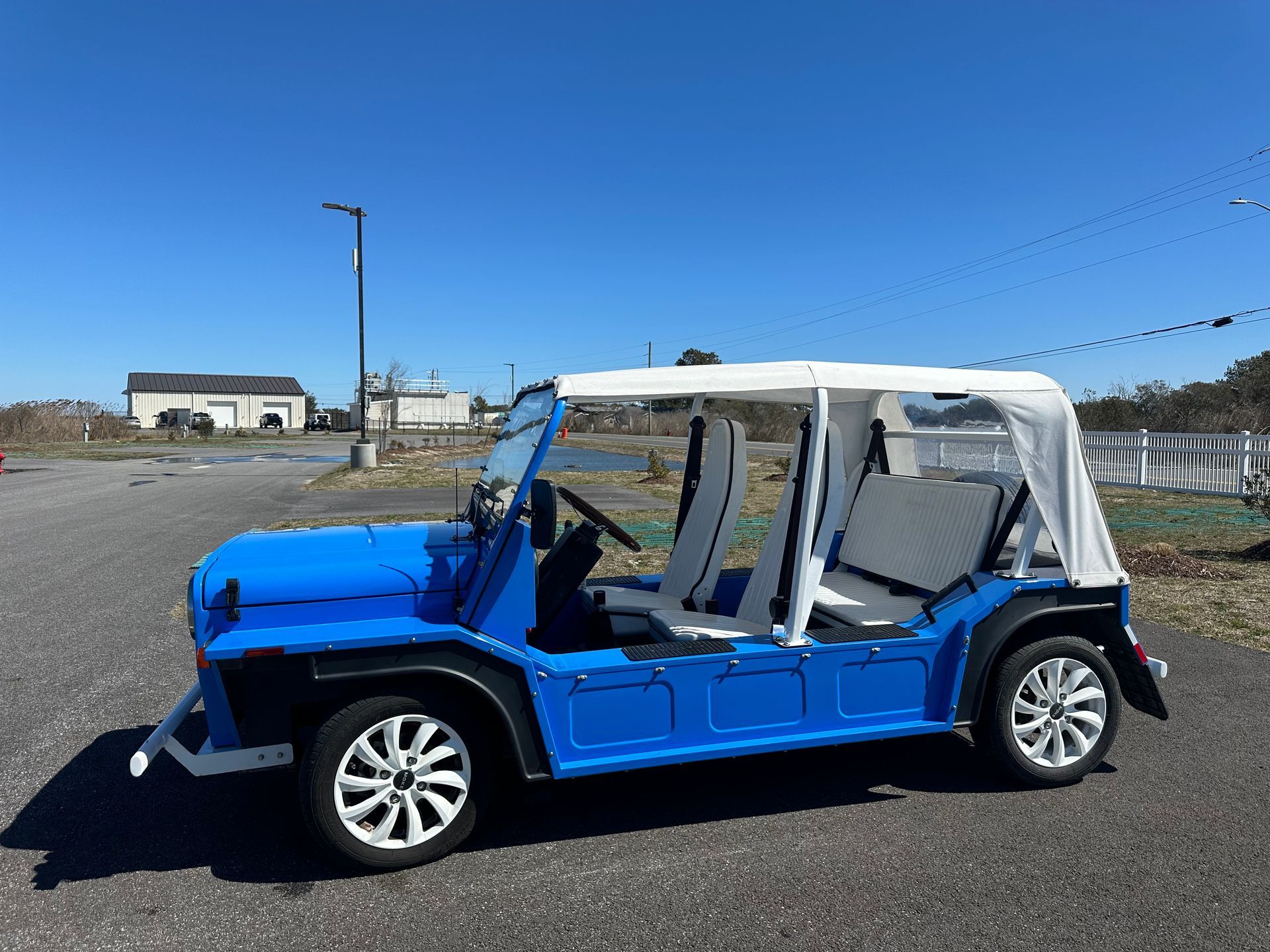 Blue Moke-style electric car with white top and wheels parked outdoors on a sunny day.