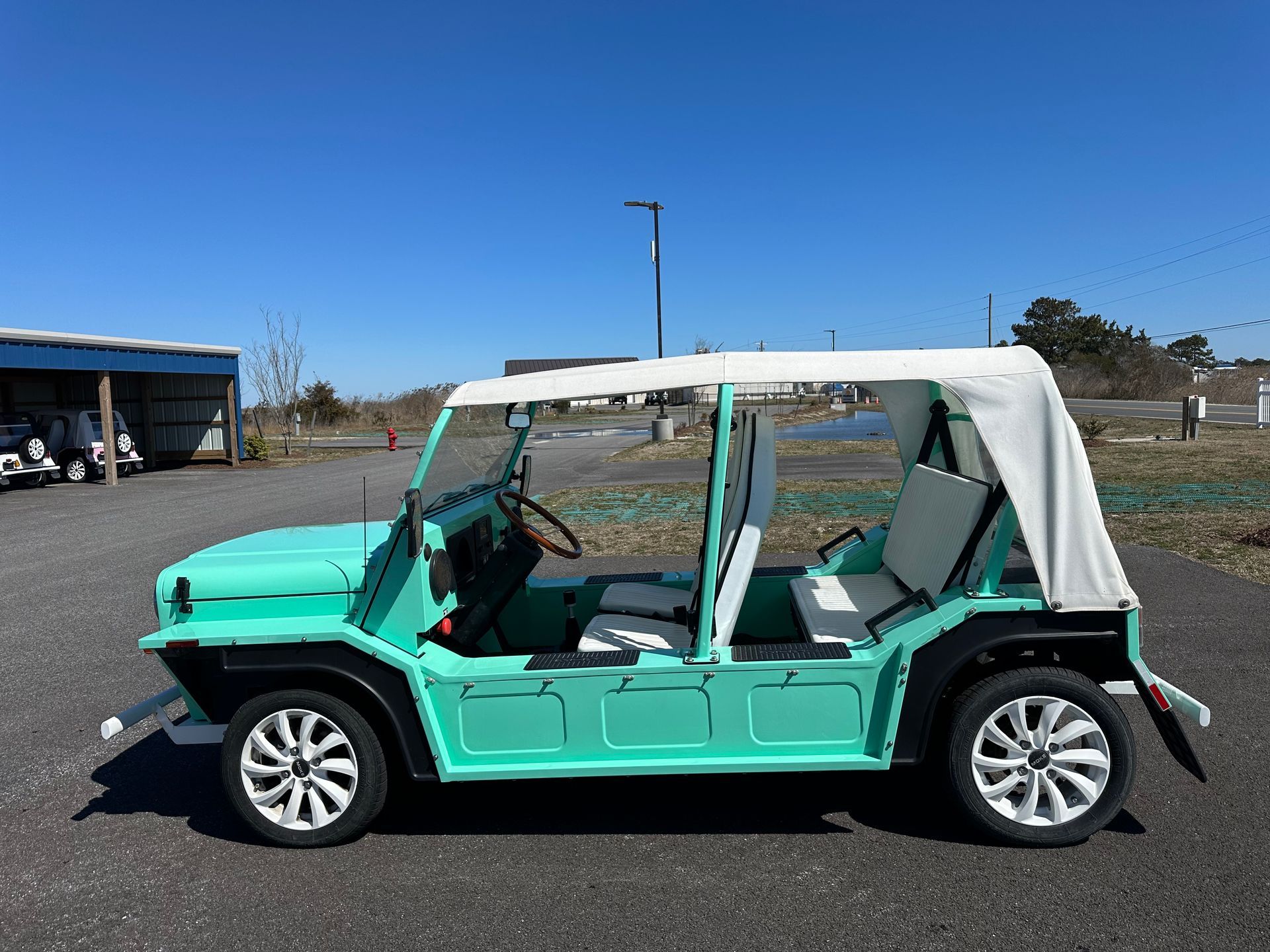 Light green Moke car with white soft top, parked outdoors on a sunny day.