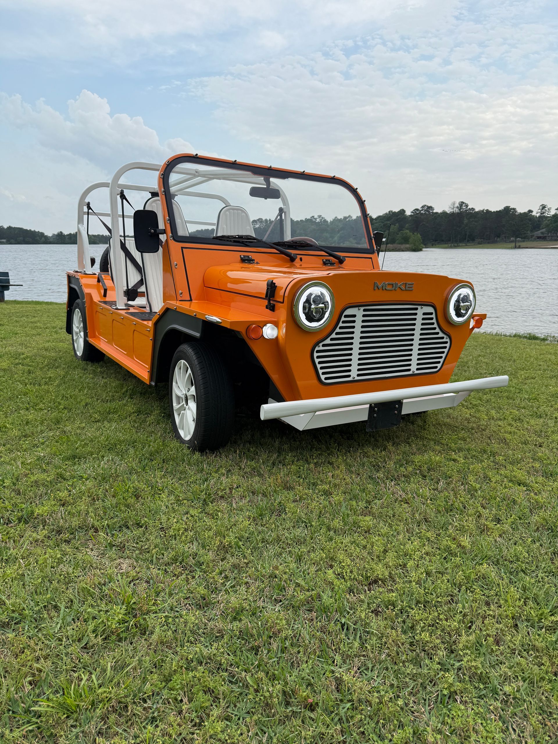 Orange open-top vehicle parked on grass near water with white roll bars and wheels.