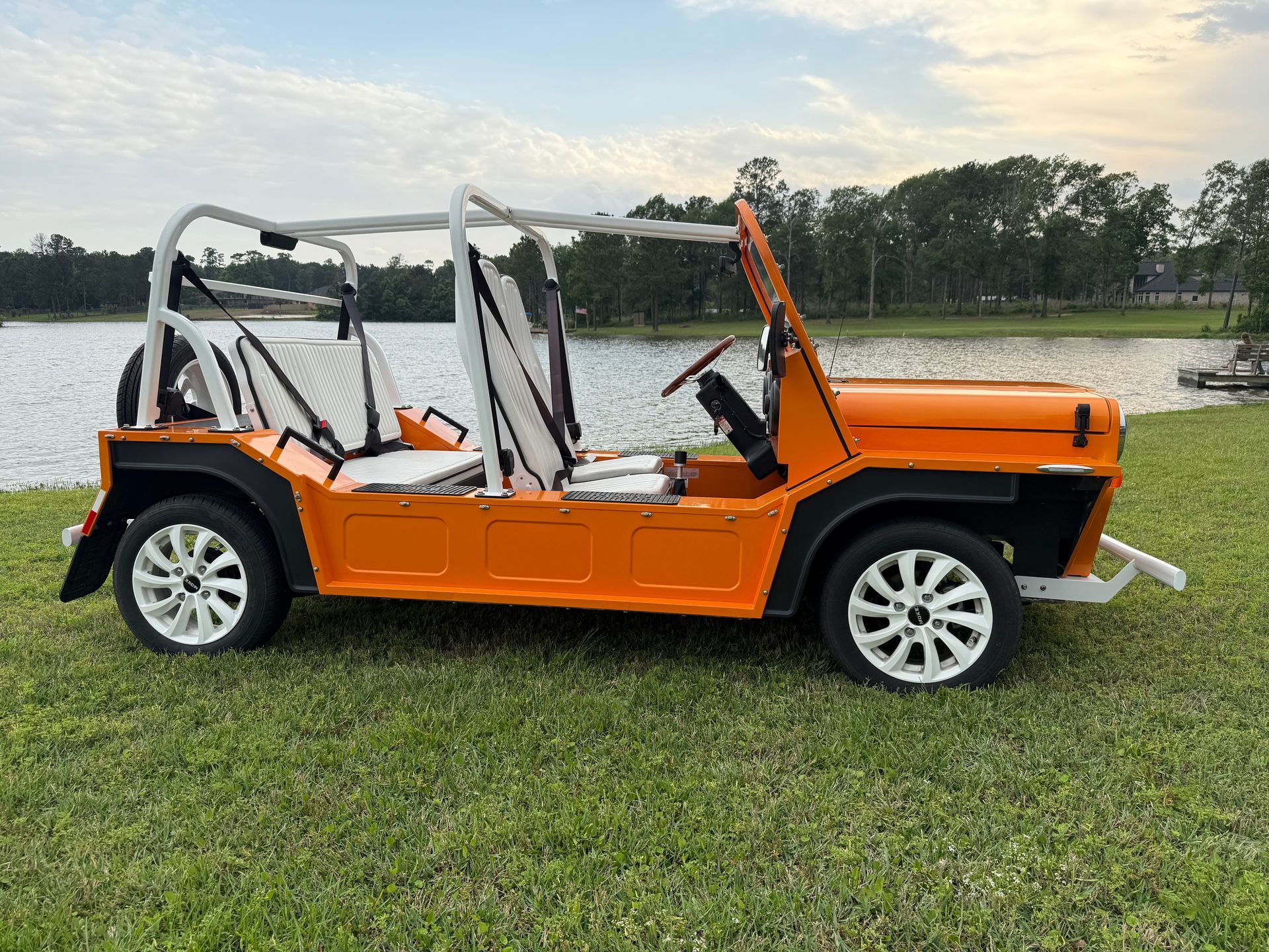 Orange open-top vehicle parked on grass near water with white roll bars and wheels.