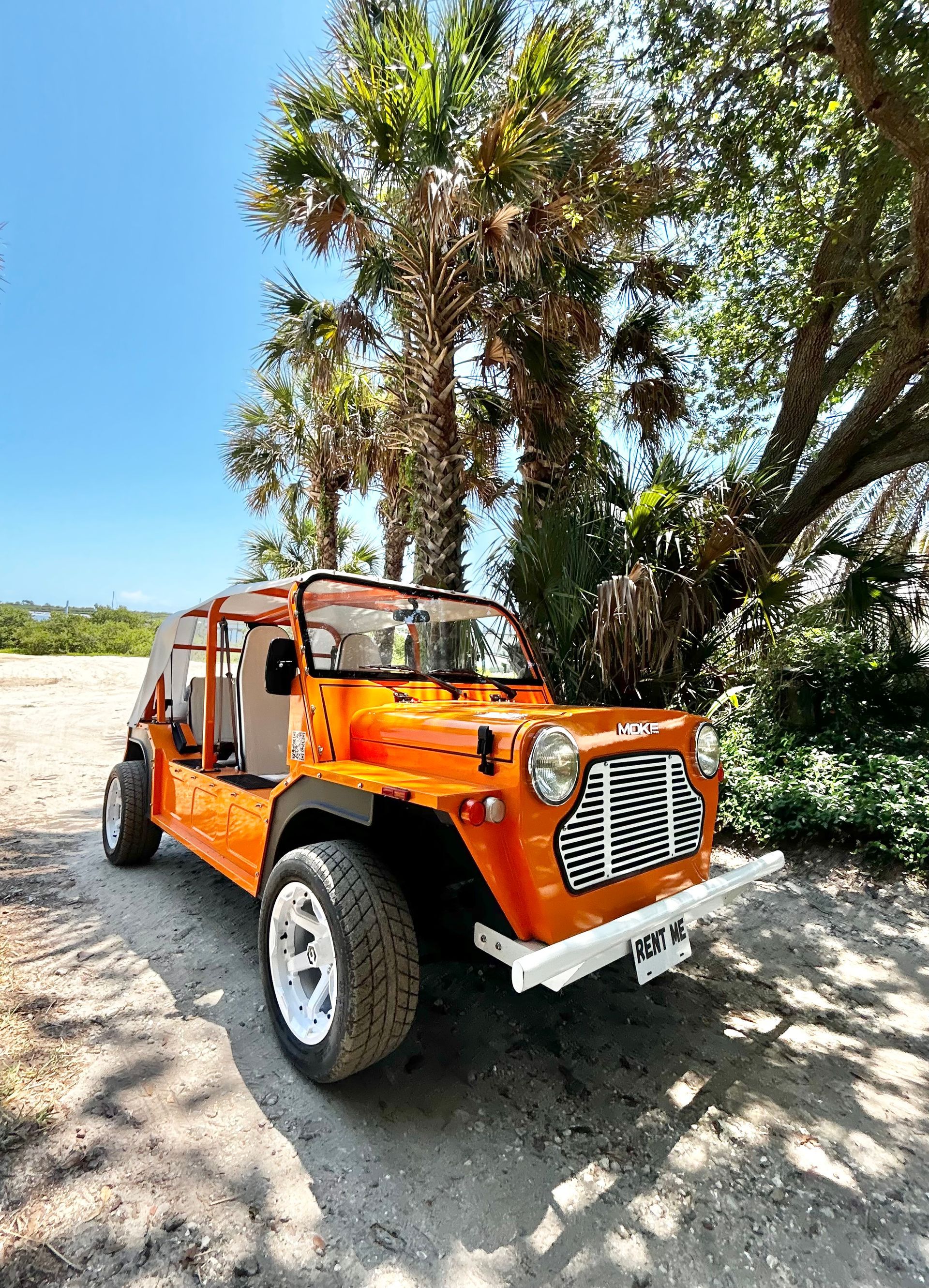Orange open-top off-road vehicle on a sandy path, white wheels, near palm trees under a blue sky.