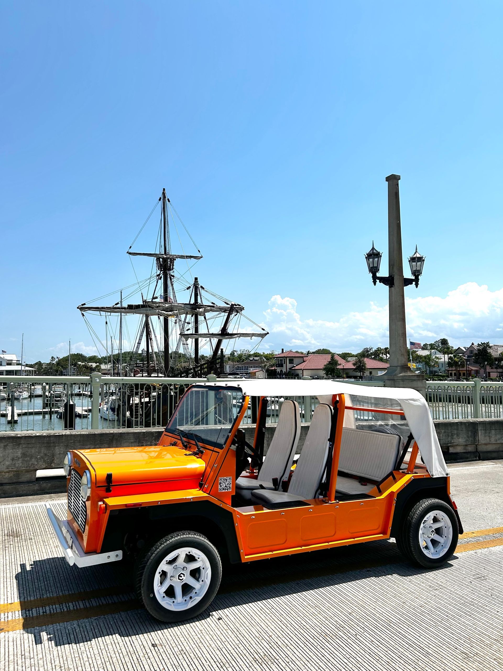 Orange open-top off-road vehicle on a sandy path, white wheels, near palm trees under a blue sky.
