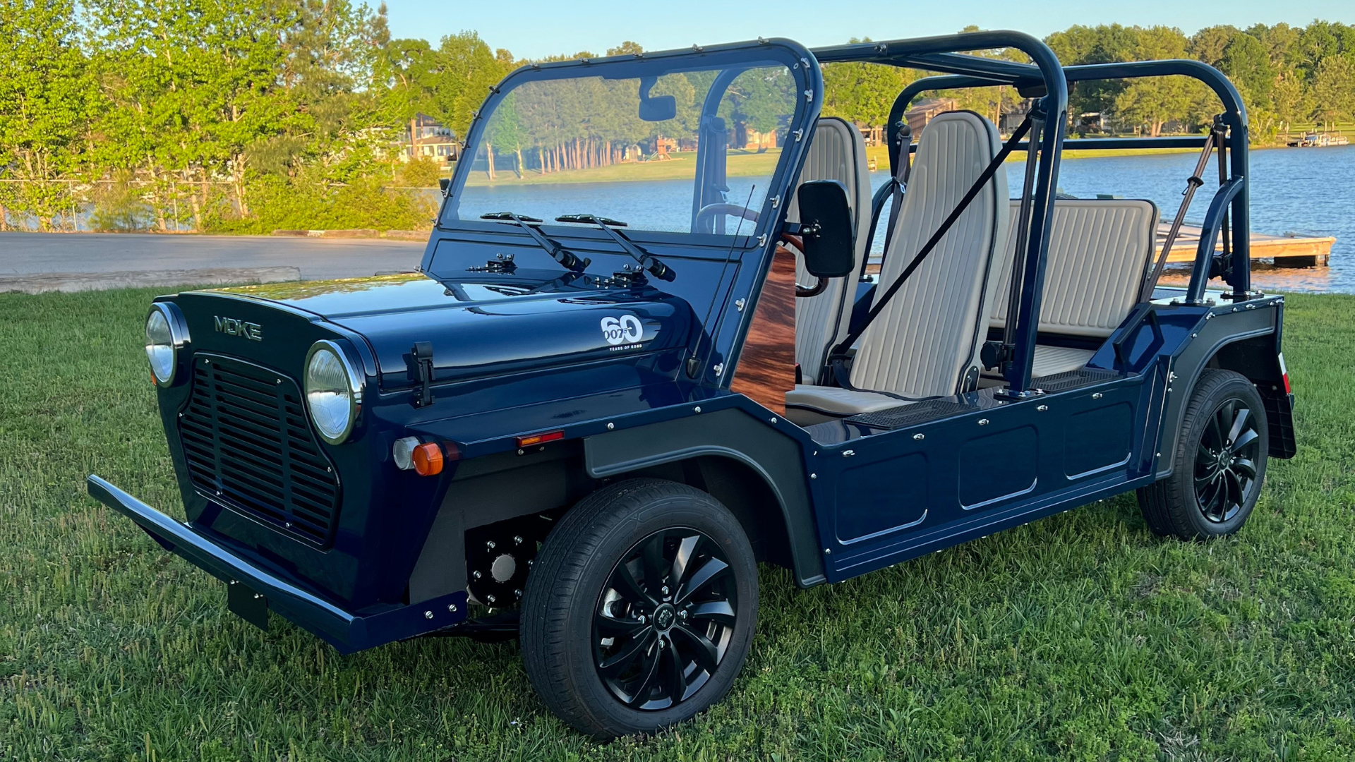 Dark blue Moke vehicle parked on grass near water. Black wheels, roll cage, tan seats.