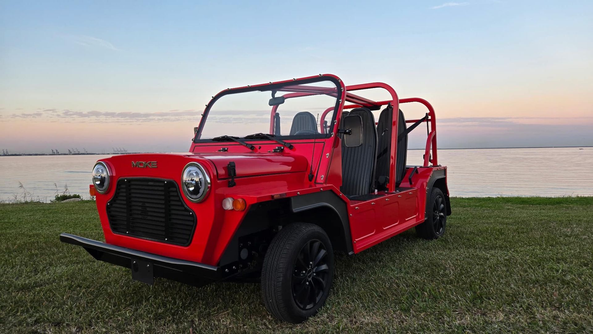 Red Moke-style open-top vehicle parked on grass near water. Black wheels, roll cage, and front bumper.