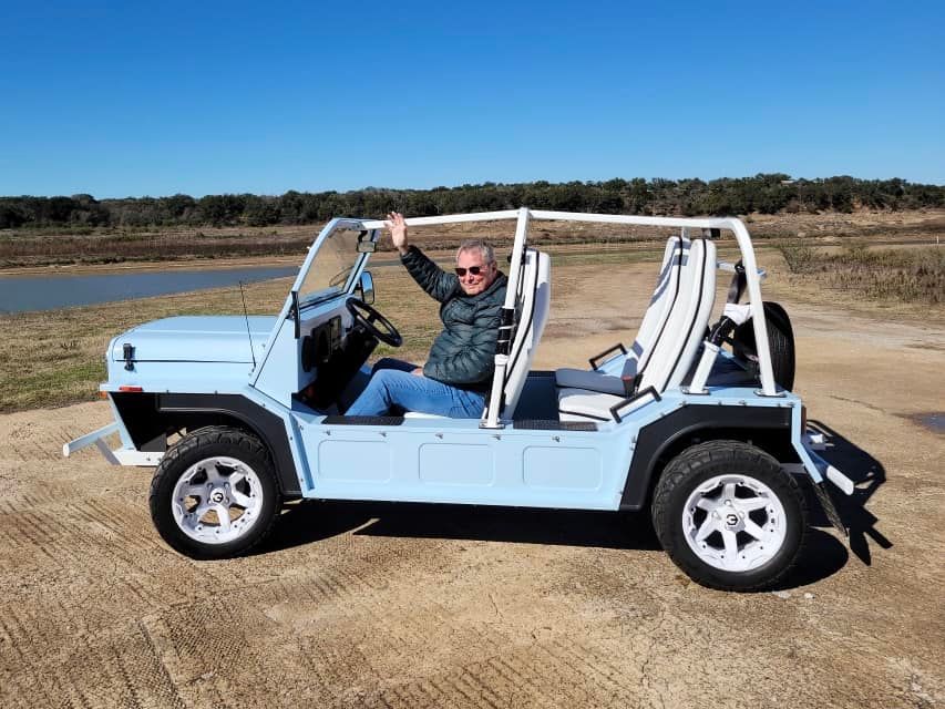Man sitting in a light blue open-top car with white roll bars, smiling, arm raised, outdoors.