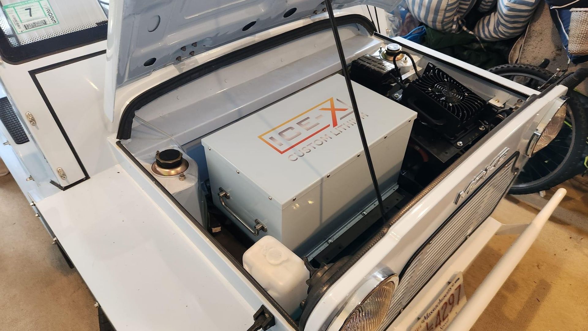 White car's open hood reveals a large, white box with an orange cross logo. Several components are visible around it.