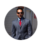 Man in a dark suit, red tie, and sunglasses standing outdoors in front of a building