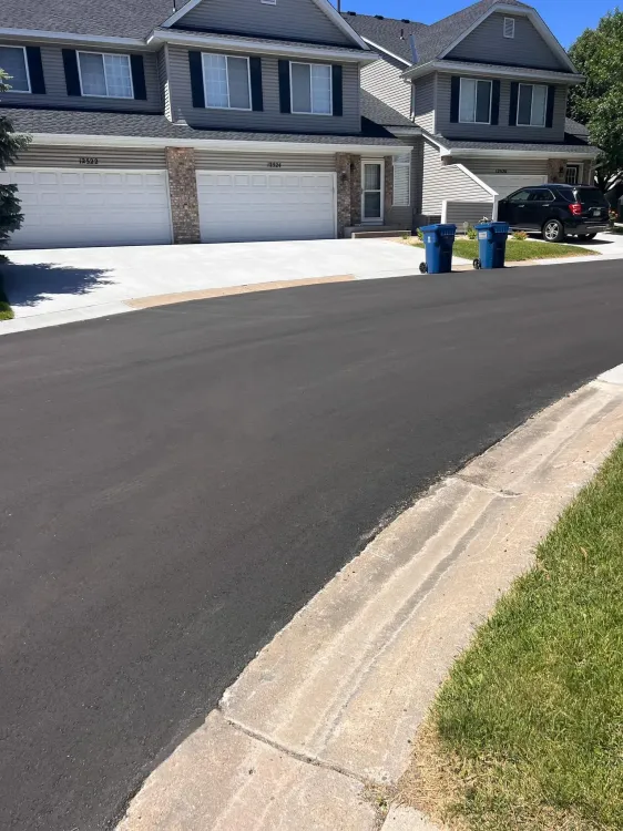 A paved street leads toward a multi-unit townhouse with attached garages, two blue trash bins, and a parked black car.