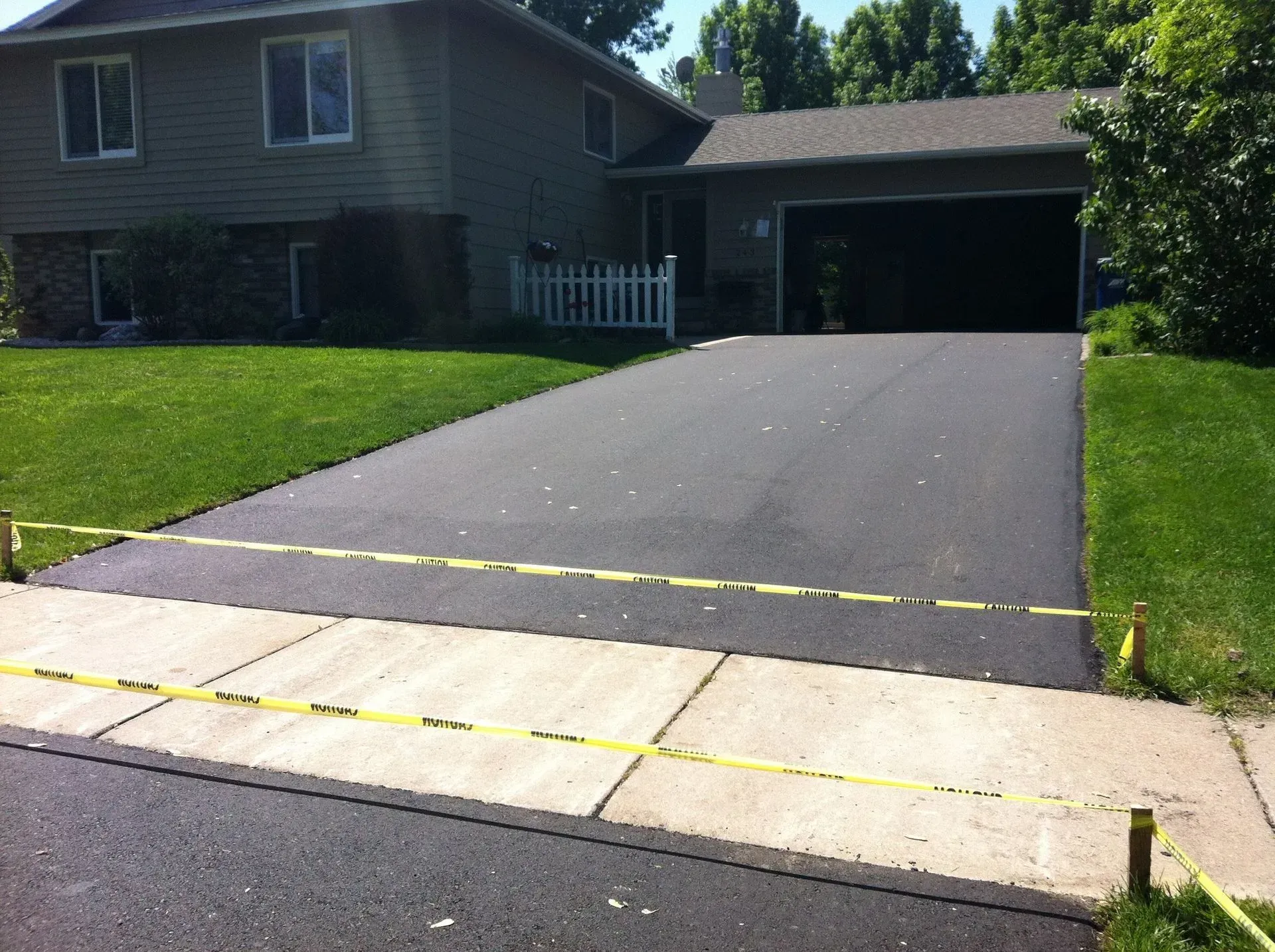 Freshly paved asphalt driveway in front of a two-story house, with caution tape across the entrance.