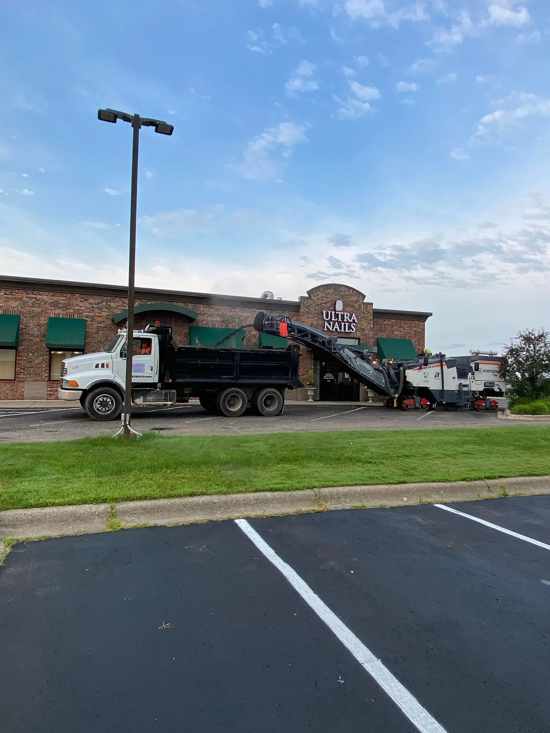 Truck loading pavement grindings from a milling machine in front of a restaurant on a sunny day.