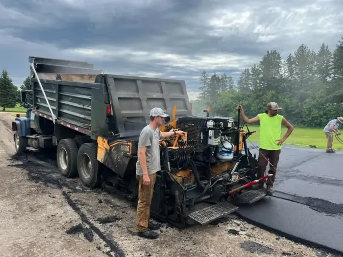 Two workers operate a black asphalt paving machine next to a dump truck on a rural road under a cloudy sky.