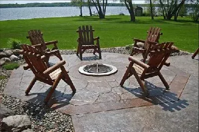 Six wooden chairs arranged around a fire pit on a patio overlooking a lake.