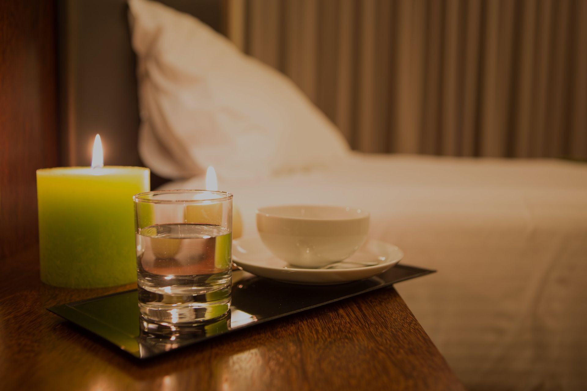 Auf einem Nachttisch neben einem Bett mit sanftem, warmem Licht stehen eine brennende grüne Kerze, ein Glas Wasser