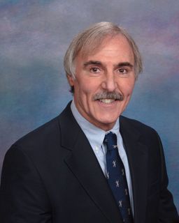 A person with a mustache smiling in a navy blazer, light blue shirt, and patterned tie against a blue backdrop.