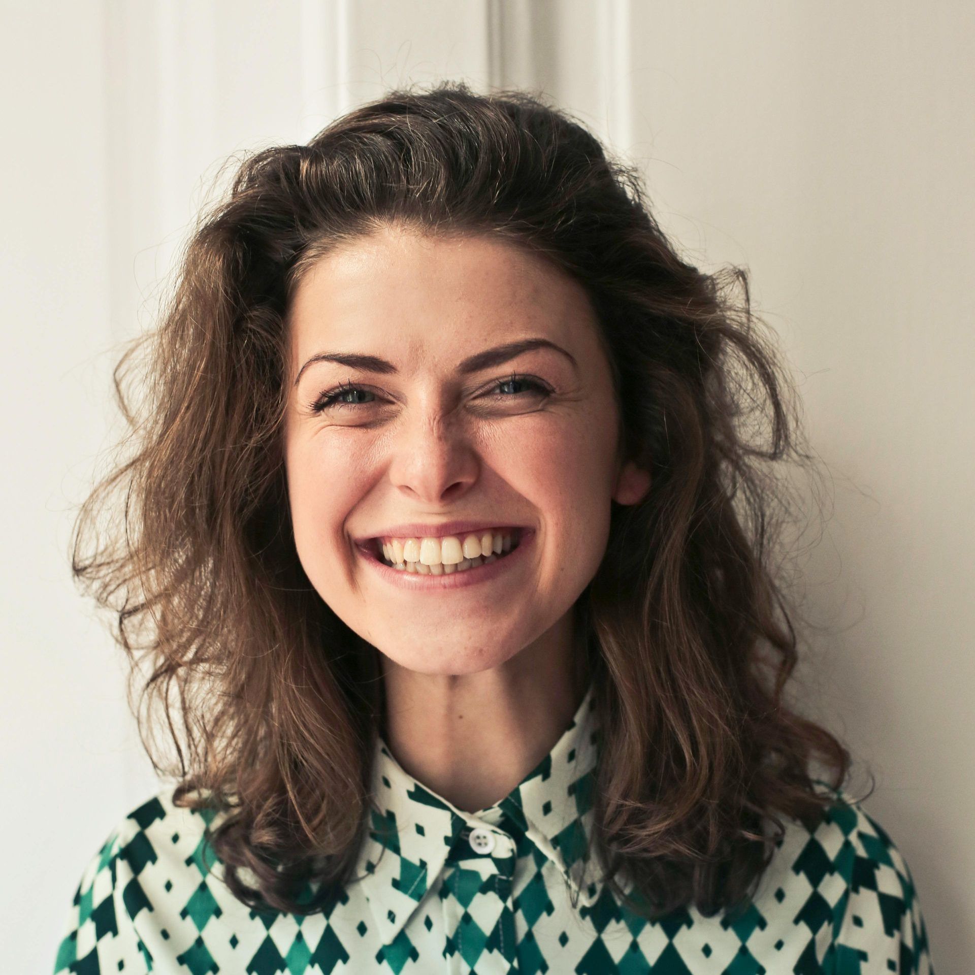 A smiling person with wavy brown hair wearing a green and white diamond-patterned collared shirt against a white wall.