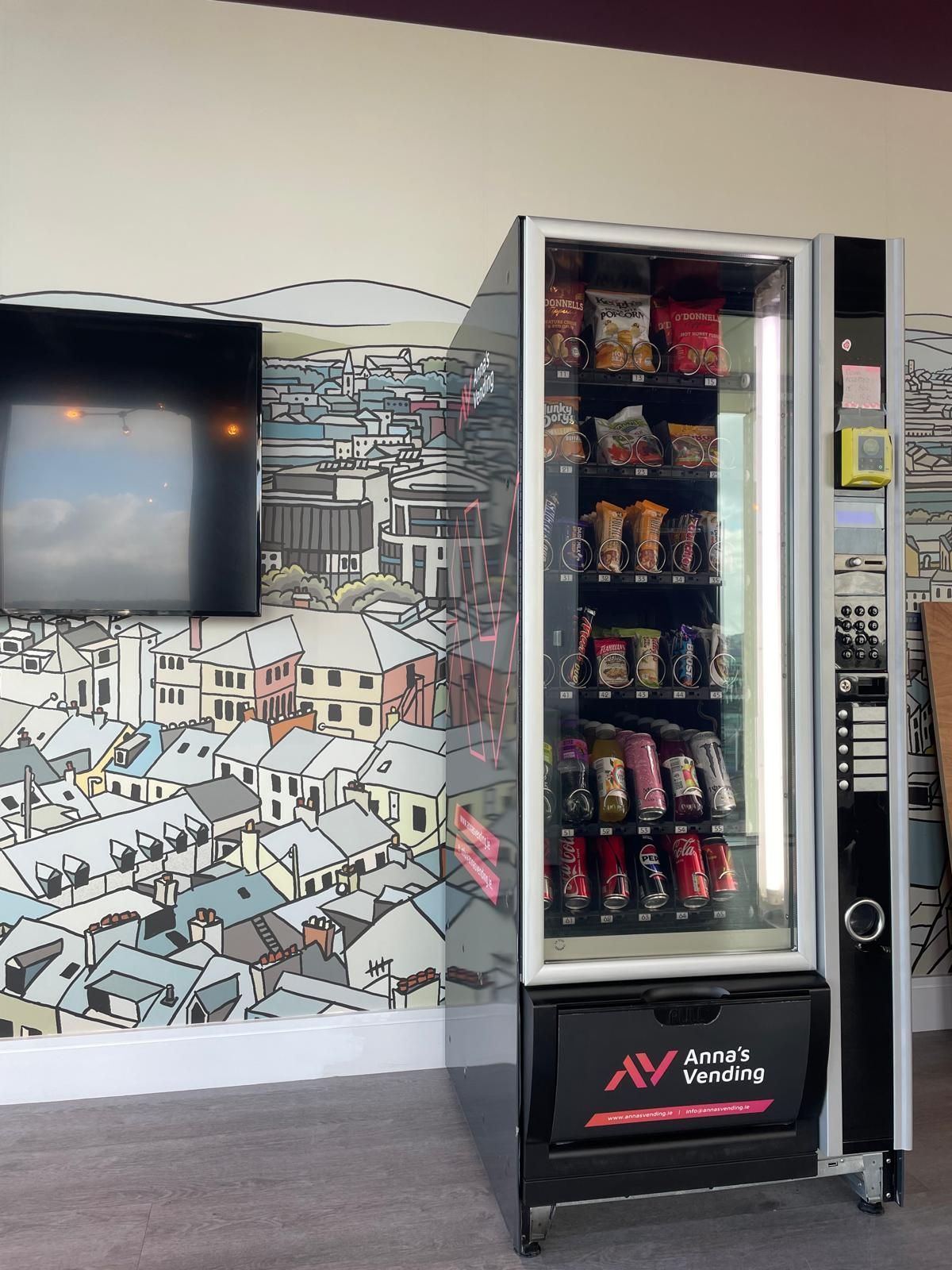 Snack vending machine in an office hallway beside a wall mural and TV screen