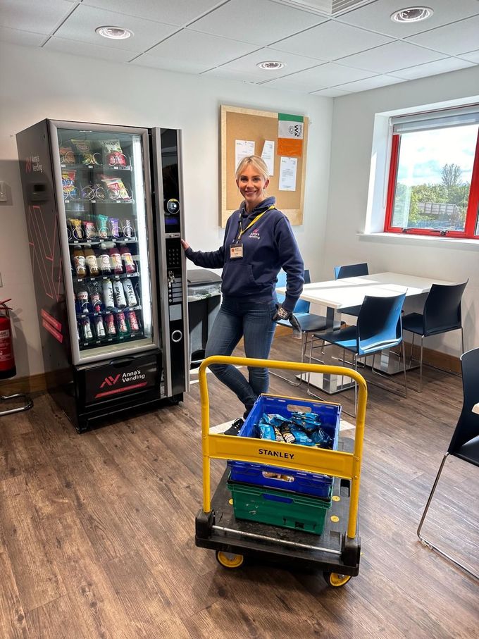 Worker beside vending machine in an office room, with a yellow utility cart in the foreground.