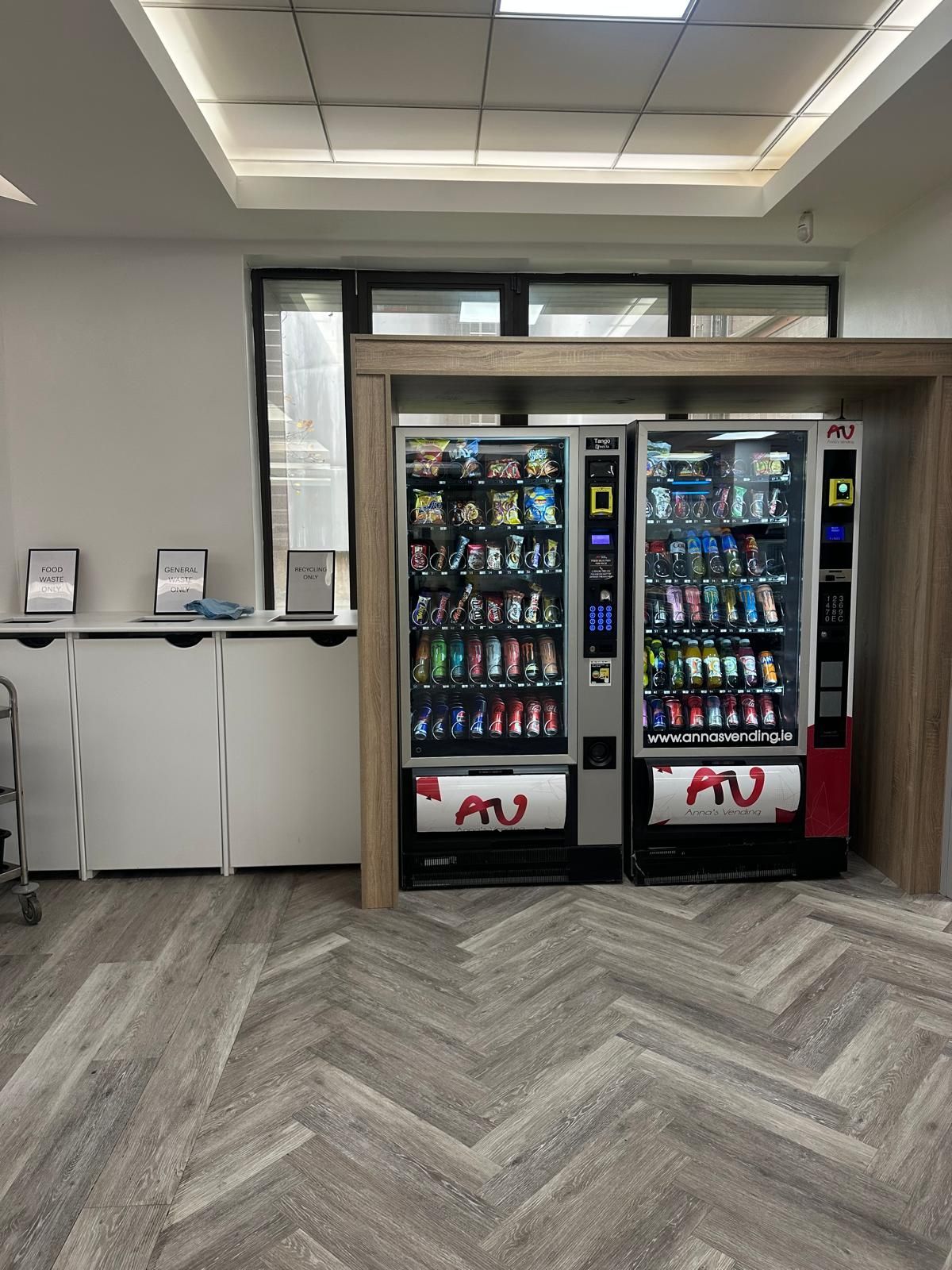 Break room with two beverage vending machines on a herringbone wood floor under fluorescent lights