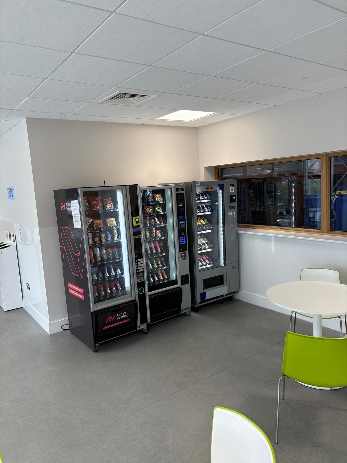 Office break room with vending machines, white walls, gray floor, and a small table with chairs.