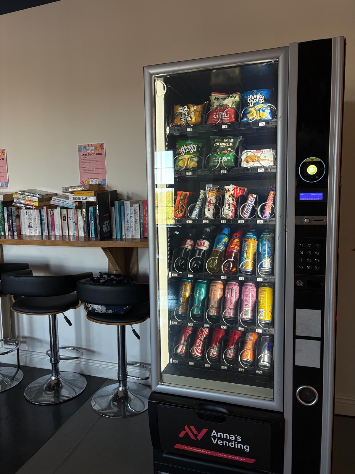 Snack vending machine beside stools and a bookshelf in a lounge area.