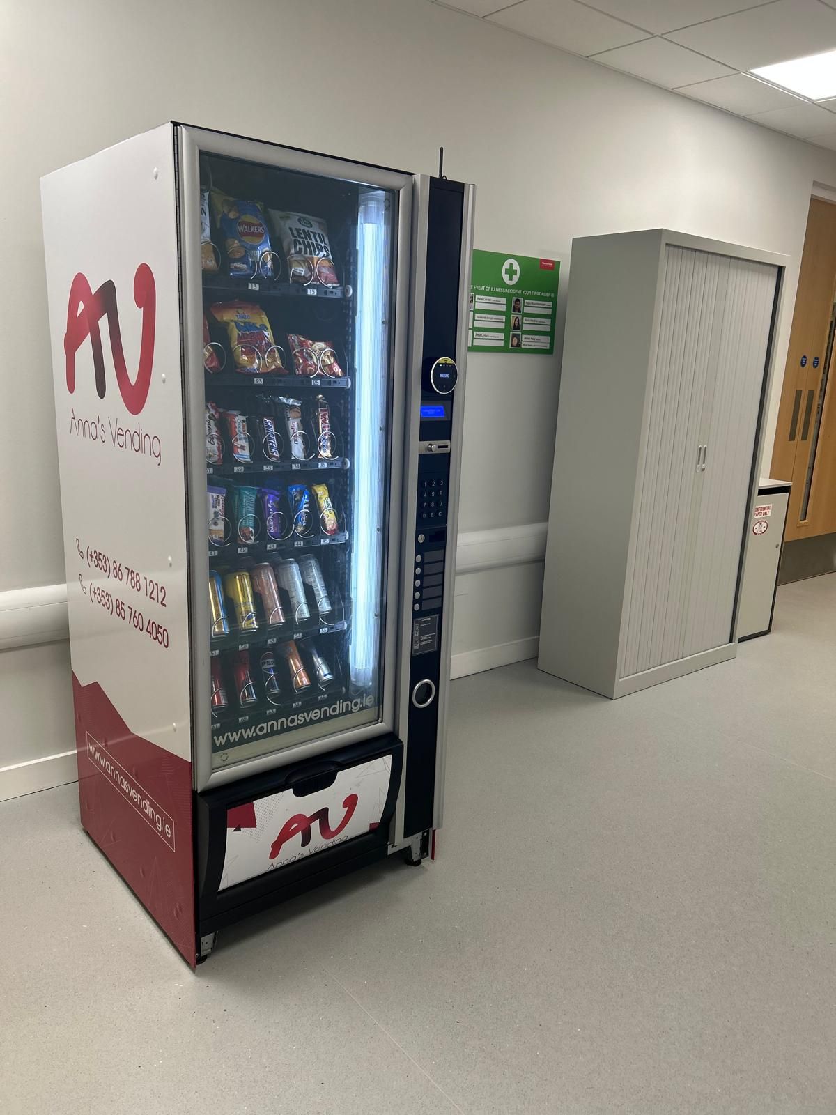 Red AV snack vending machine in a hallway beside beige lockers.