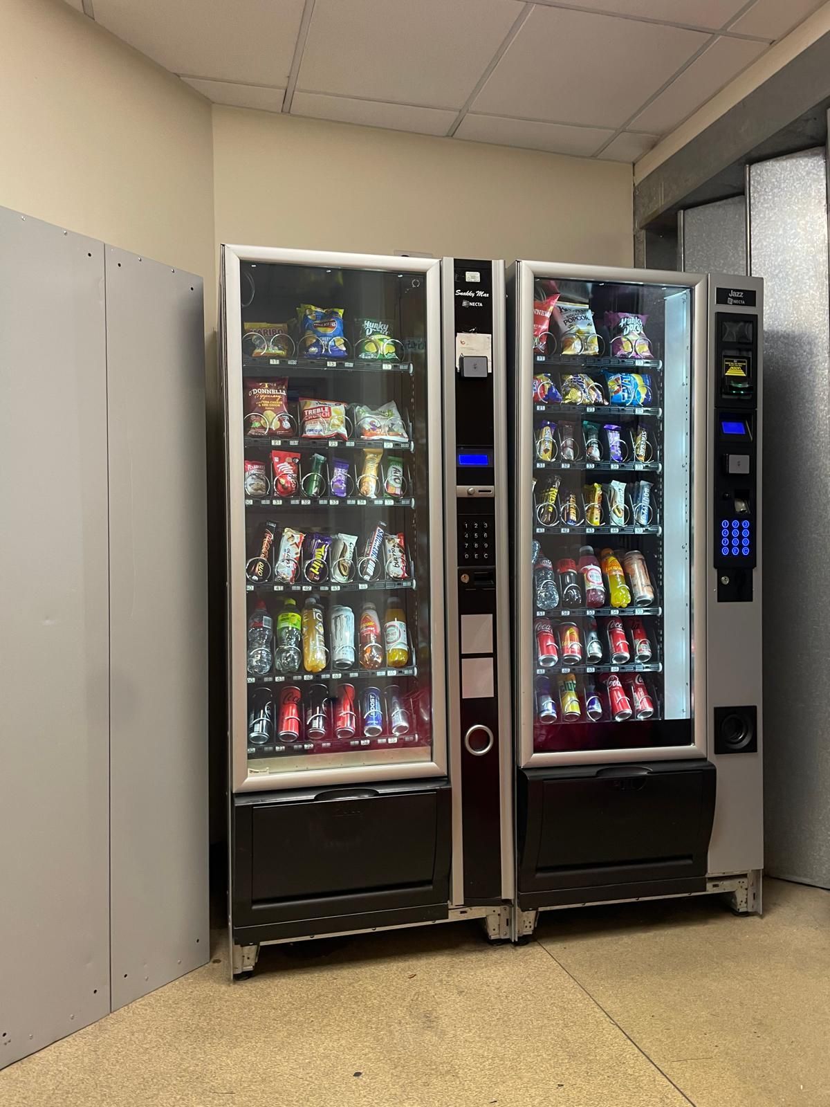 Two vending machines with snacks and drinks in a beige hallway, one lit display in front of a wall cabinet