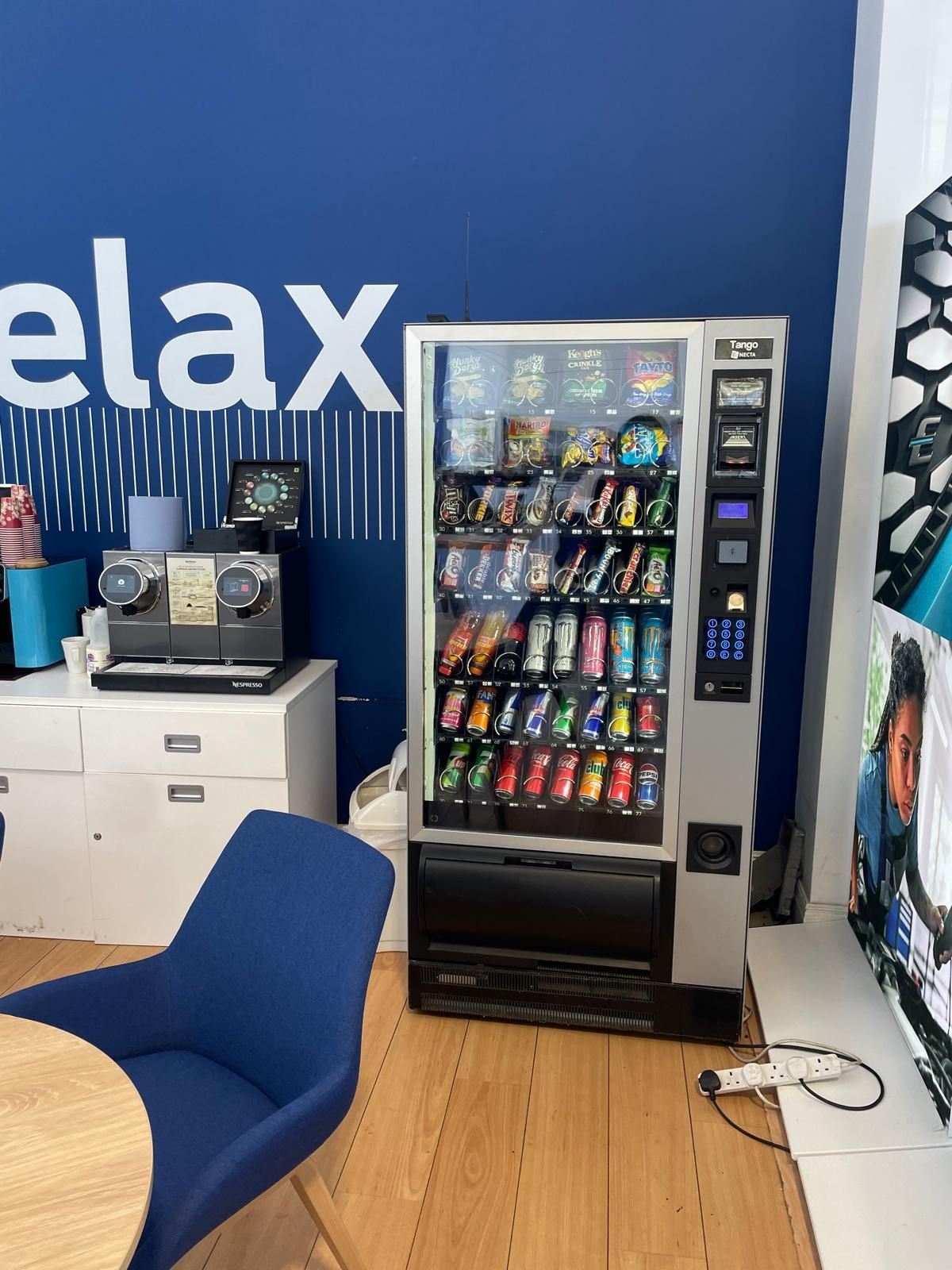 Office break room with blue wall, snack vending machine, and chairs beside a small table.