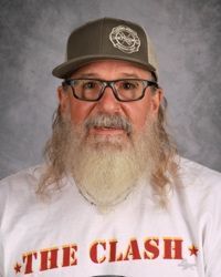 Man with glasses, gray beard, and cap, wearing a white “THE CLASH” shirt against a gray backdrop