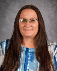 Portrait of a woman with glasses and long brown hair, wearing a blue patterned top against a gray backdrop