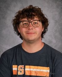 Person with curly hair and glasses wearing a dark T-shirt against a gray studio backdrop