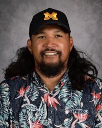 Smiling person in a black cap with a yellow M, wearing a dark floral shirt against a gray backdrop