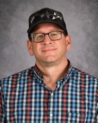 Portrait of a man wearing glasses, a black cap, and a blue-red plaid shirt on a gray background