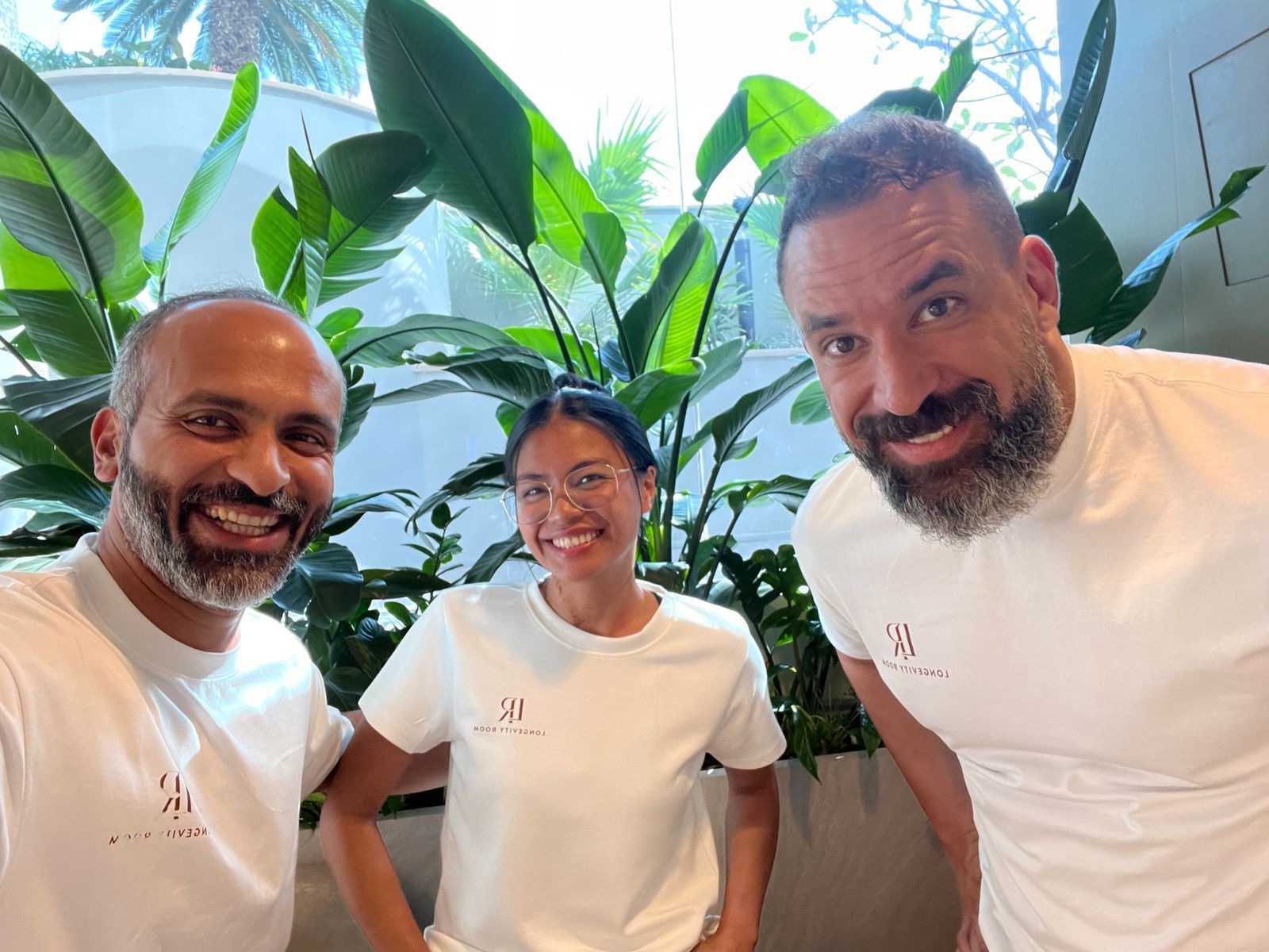 Three people smiling indoors among large green plants, wearing white shirts.