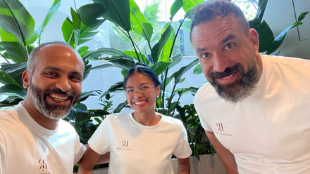 Three smiling people in white shirts posing indoors with large green plants behind them