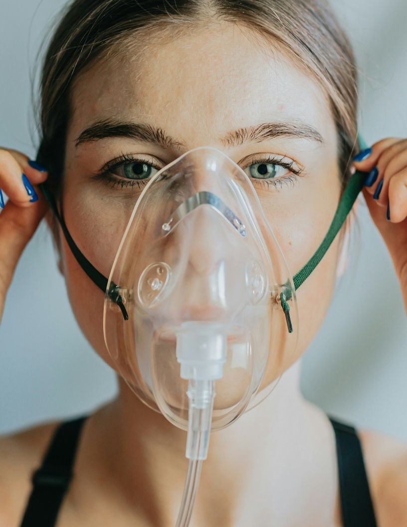 Person adjusting a clear oxygen mask, looking at the camera against a plain background